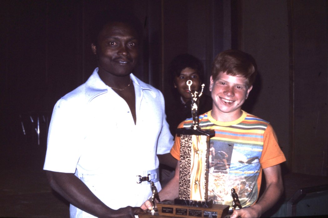A young athlete in an orange t-shirt is shaking hands with a man wearing a white t-shirt awarding him with a trophy. They are both smiling and posing for the camera.