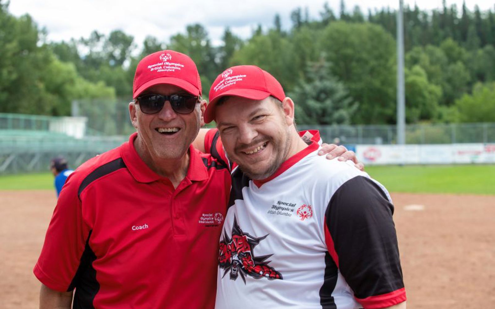 An athlete and coach wearing baseball caps smile for a photo together.