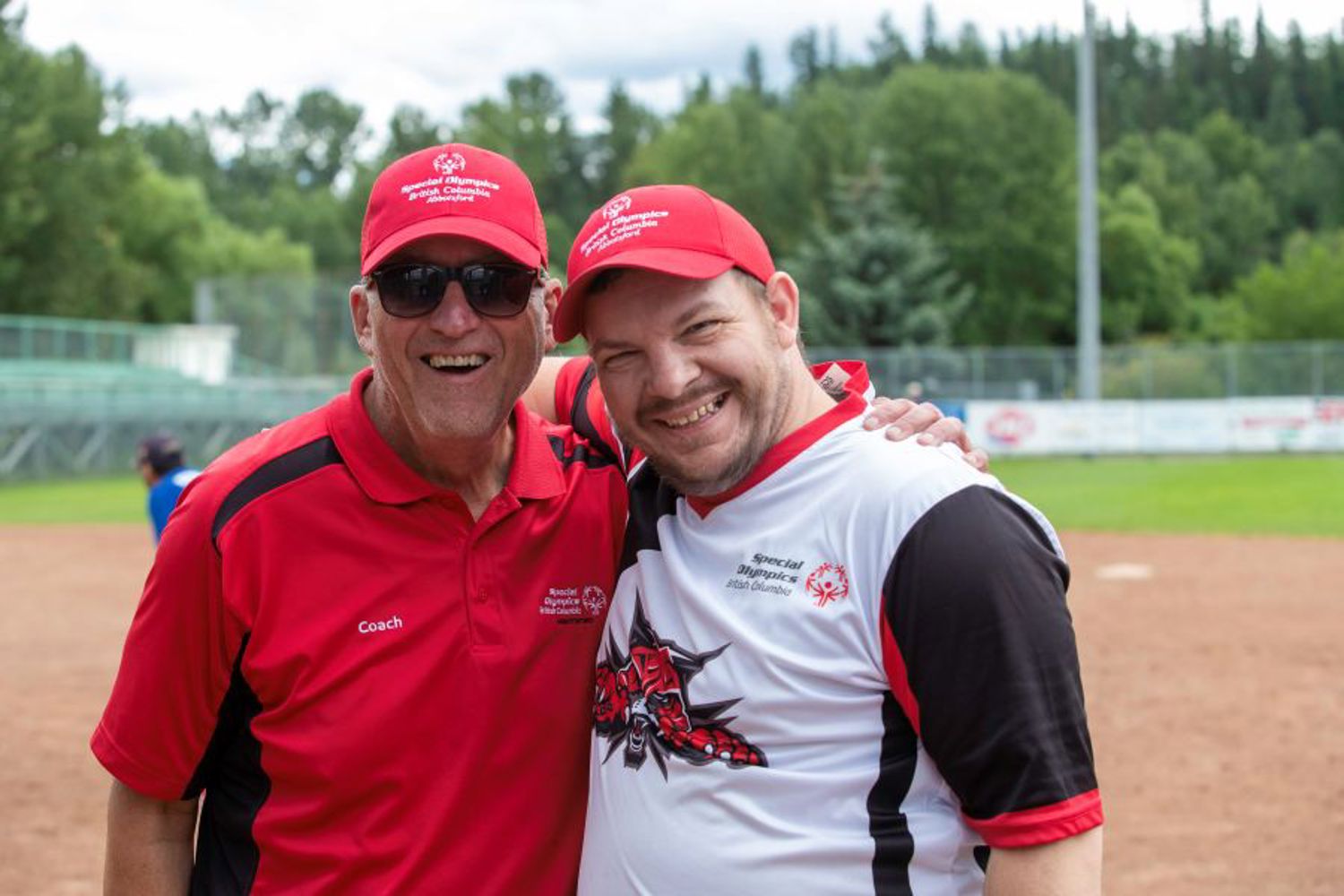 An athlete and coach wearing baseball caps smile for a photo together.