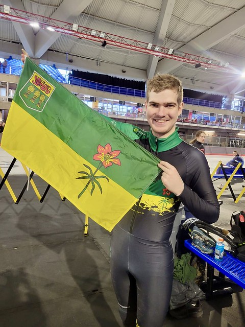 A picture of Jordan, a Special Olympics Saskatchewan athlete holding up a Saskatchewan flag. He is standing in an ice rink but next to the ice. A picture of Jordan, a Special Olympics Saskatchewan athlete holding up a Saskatchewan flag. He is standing in an ice rink but next to the ice.
