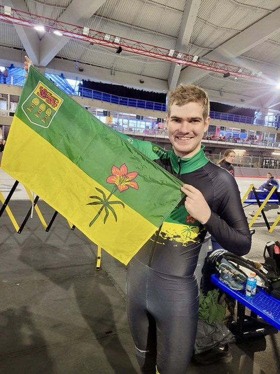 A picture of Jordan, a Special Olympics Saskatchewan athlete holding up a Saskatchewan flag. He is standing in an ice rink but next to the ice.