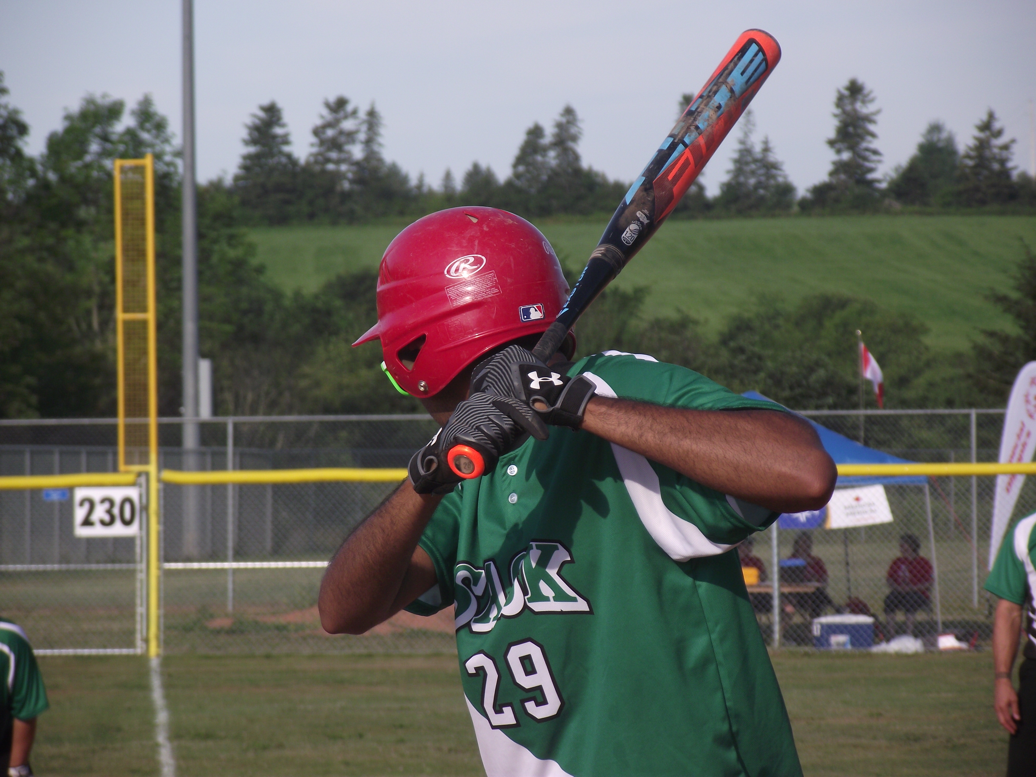 A close up image of an athlete playing baseball. They have their face turned away from the camera as they are focusing on their game. They are swinging the baseball bat back in preparation to hit the ball. They are wearing a green baseball jersey. A close up image of an athlete playing baseball. They have their face turned away from the camera as they are focusing on their game. They are swinging the baseball bat back in preparation to hit the ball. They are wearing a green baseball jersey.