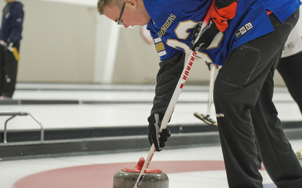 A close up image of a curling athlete. The athlete is holding his curling stick in front of the curling stone. He is wearing a blue jersey.