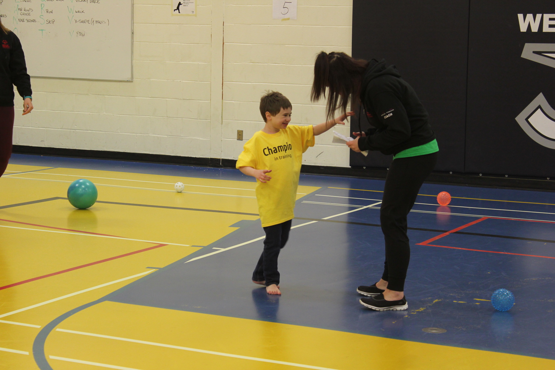 A young athlete in a yellow t-shirt that says "Champion in Training" on it, is reaching his arm out towards his instructor. They are both in a gymnasium and different coloured balls are scattered across the floor.