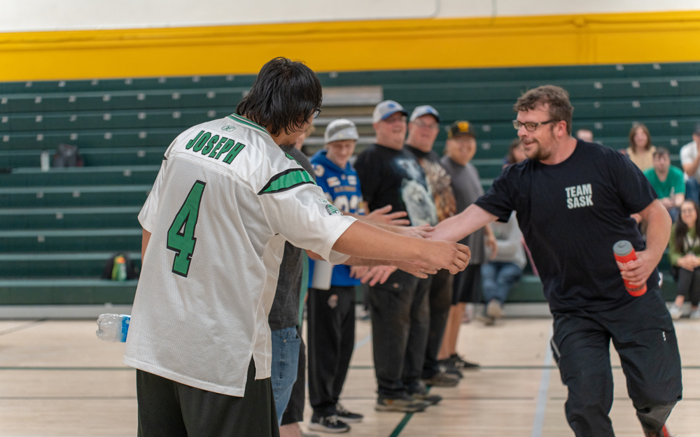 A man wearing a black t-shirt that says "Team Sask" on it, is walking down a lineup of athletes and giving them a high-five. The athlete in the front of the line is in the focus of the camera. He is wearing a white and green jersey with "Joseph" and the number "4" on the back. Everyone is in a gymnasium.