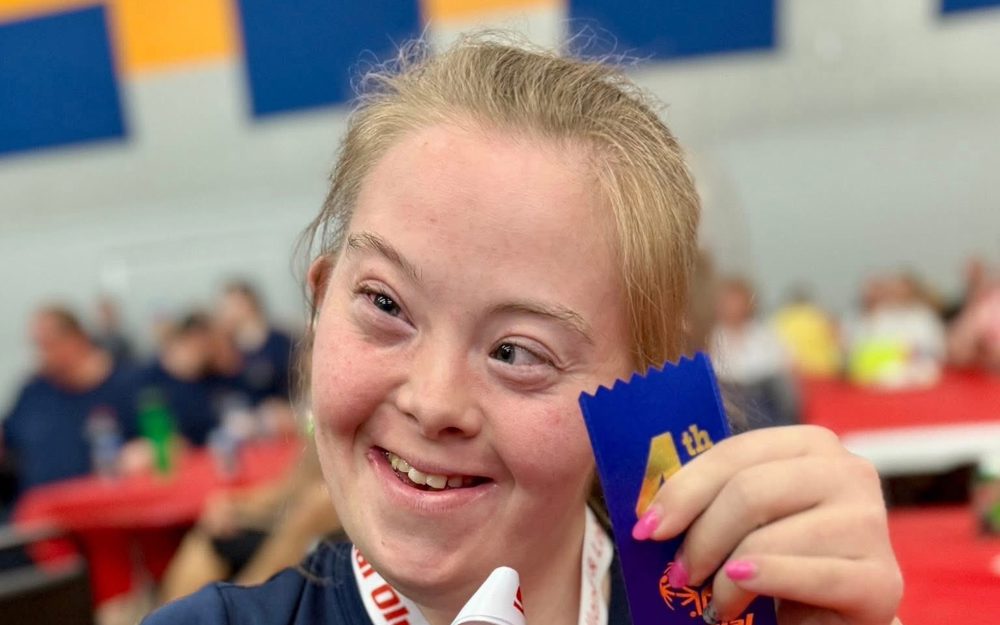 A close up image of an athlete holding up the medal and ribbon she won. The silver medal is hanging around her neck, and she is holding up the medal in one hand. In the other hand is a blue 4th place ribbon. The athlete is wearing a navy blue t-shirt with the Special Olympics Newfoundland & Labrador CBS Brightstars logo on it.