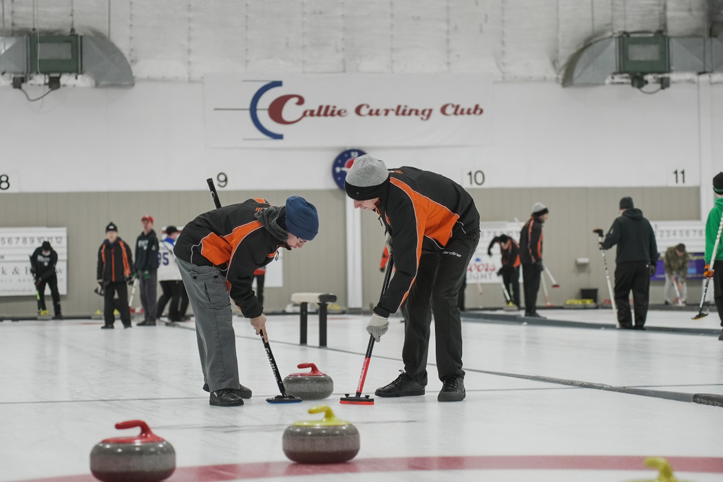 An action shot of a curling game. Two curling athletes are using their curling sticks to help the curling stone slide across the ice. They are wearing black and orange jackets and are focused on the game.