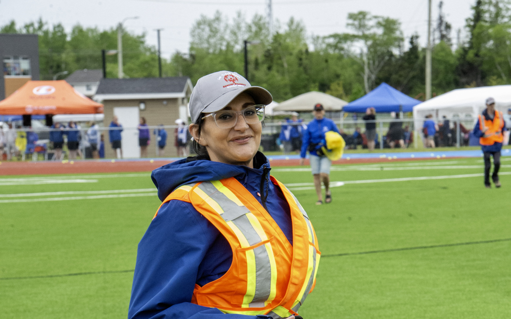 A photo of a volunteer mid run, wearing a bright orange and yellow safety vest, and a Special Olympics Newfoundland & Labrador grey baseball cap.