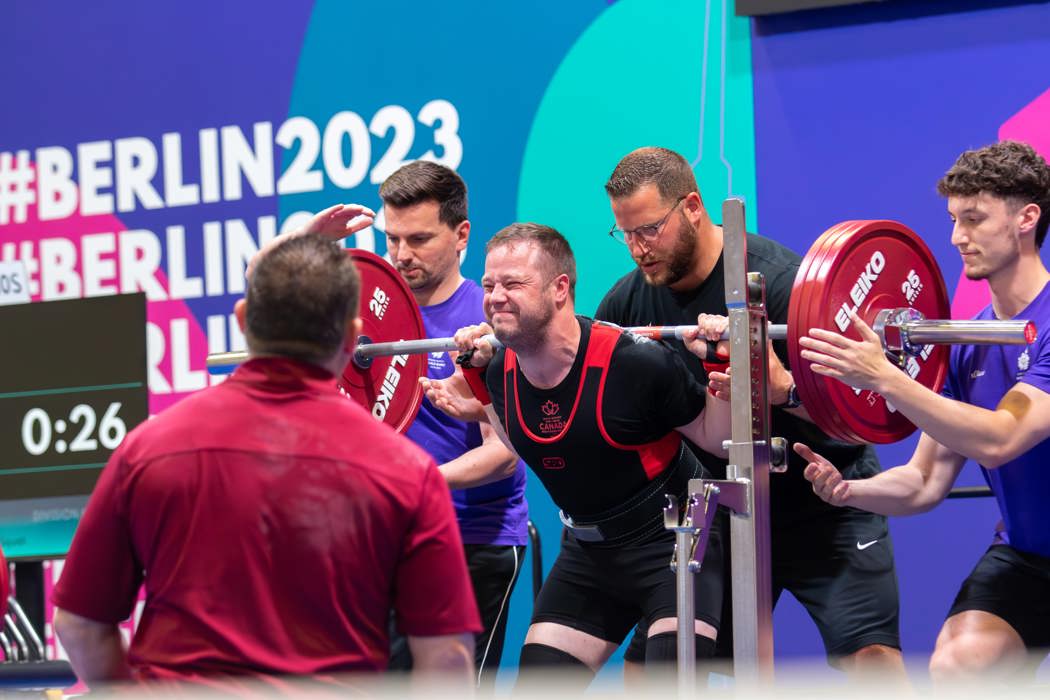 Young man gritting his teeth to lift a heavy barbell. Other people stand nearby to help as needed. 