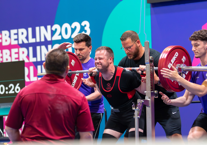 Young man gritting his teeth to lift a heavy barbell. Other people stand nearby to help as needed.