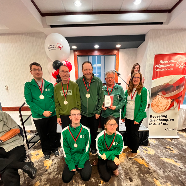 A group team photo of athletes who have won medals. They pose, two people in front on their knees and five standing behind them. They are all wearing green sweaters with a ribboned medal hanging around their necks. The image takes place in a large room with banners and balloons behind the team.