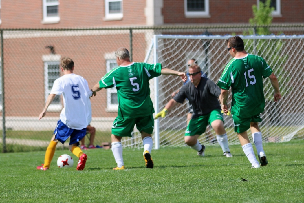 An action shot photo of a soccer match. Four athletes can be seen on the soccer field playing soccer. One player in a white jersey has possession of the soccer ball and is approaching the net. The goalie in the net has his arms out ready to guard the net. Two players in green jerseys are defending the net and running towards the player in possession of the ball.