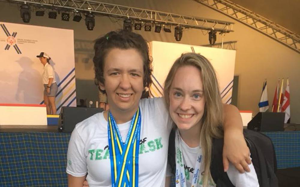 A photo of a volunteer and athlete with their arms around behind each other smiling for the camera. They are both wearing white t-shirts. The athlete has medals around her neck while the volunteer has a colourful lanyard around her neck.