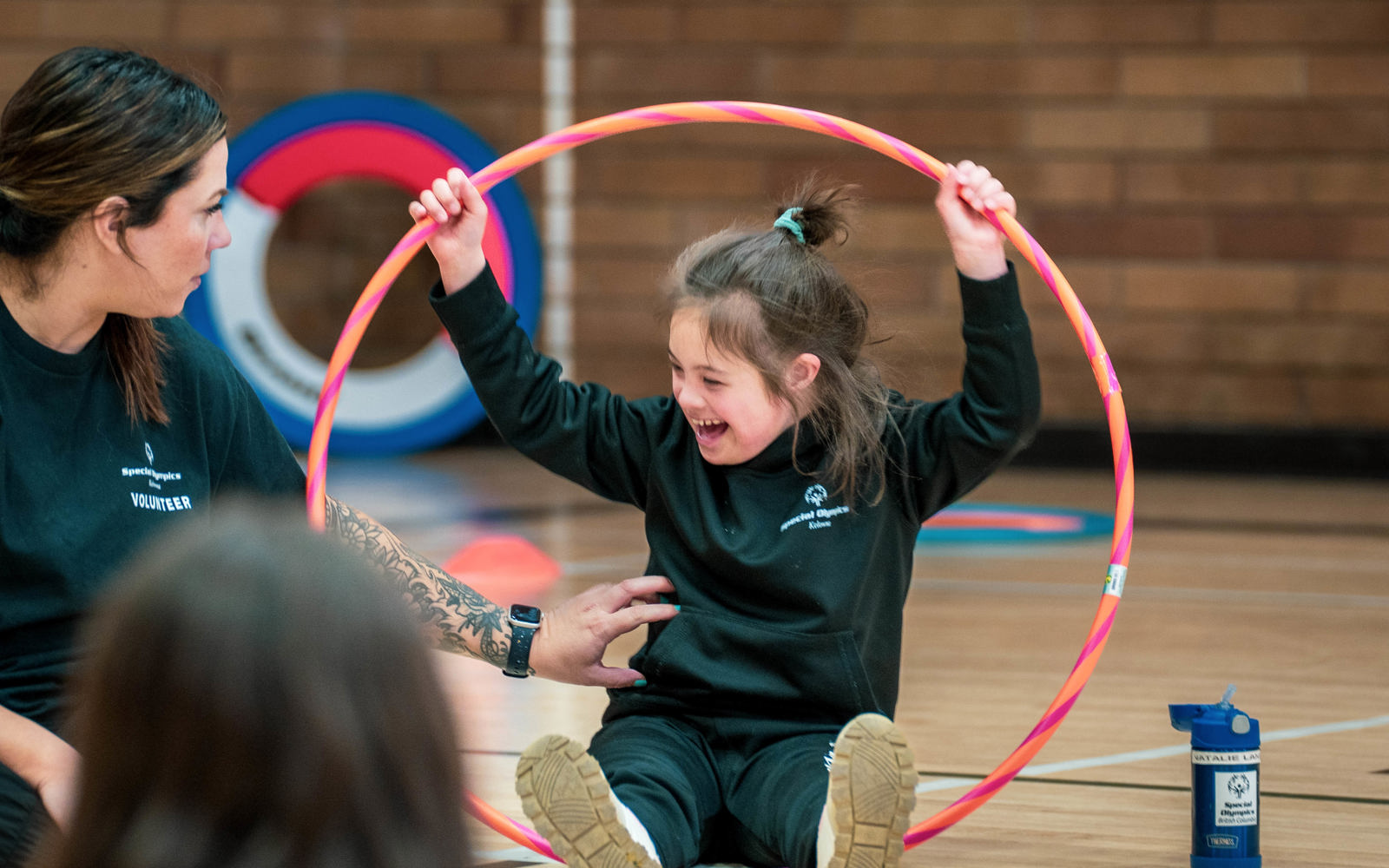 A young girl with intellectual disabilities sits on the floor of gym holding a large, pink hula-hoop over her head while interacting with her leader.