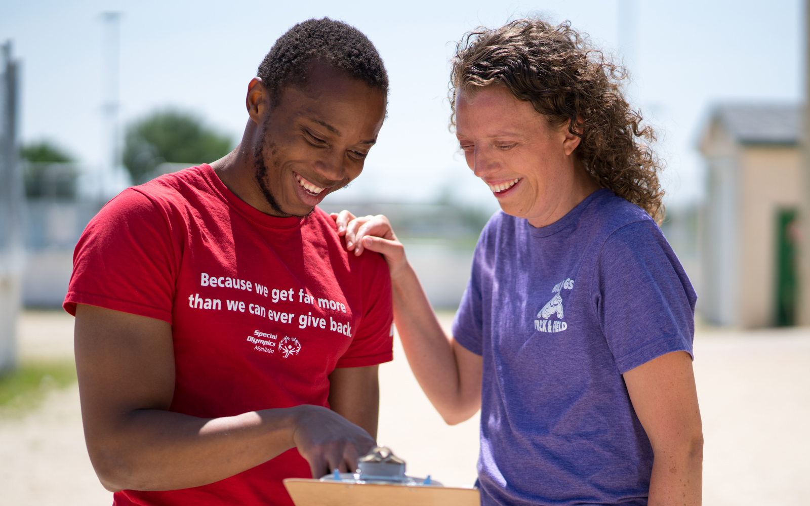 A person in a red shirt with the words "Because we get far more than we can ever give back" holding and pointing at a clipboard while a person in a purple shirt smiles down at the clipboard. 