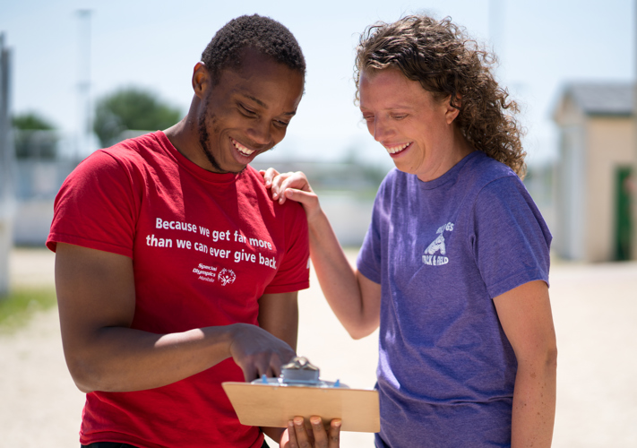 A person in a red shirt with the words "Because we get far more than we can ever give back" holding and pointing at a clipboard while a person in a purple shirt smiles down at the clipboard.