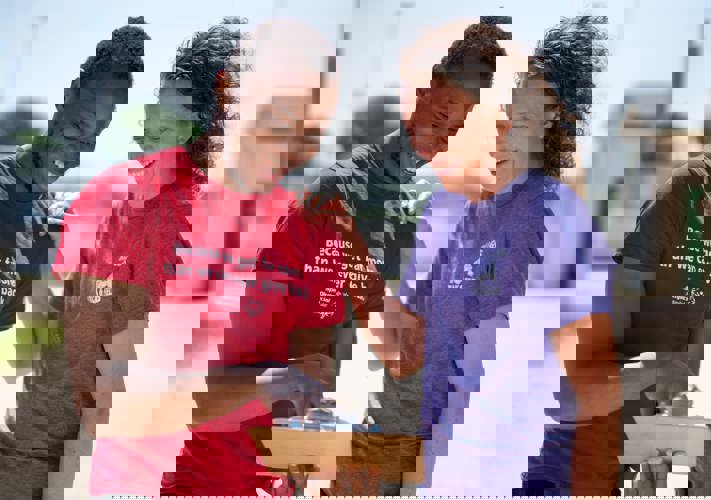 A person in a red shirt with the words "Because we get far more than we can ever give back" holding and pointing at a clipboard while a person in a purple shirt smiles down at the clipboard.