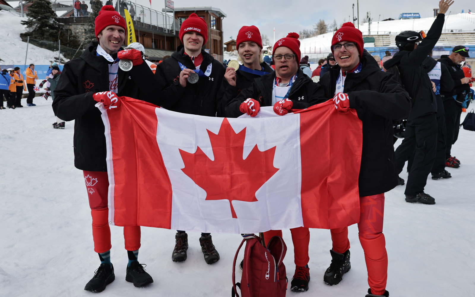 Five young men holding the Canadian flag while wearing medals around their necks. The background is outside and is covered in snow. All the men are smiling and looking happy.