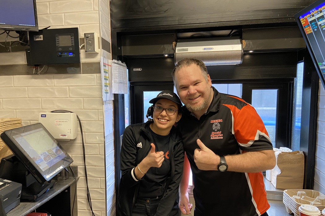 A Special Olympics Saskatchewan athlete is posing with a Tim Hortons employee at the drive through window. They are both giving a thumbs up and smiling for the camera.