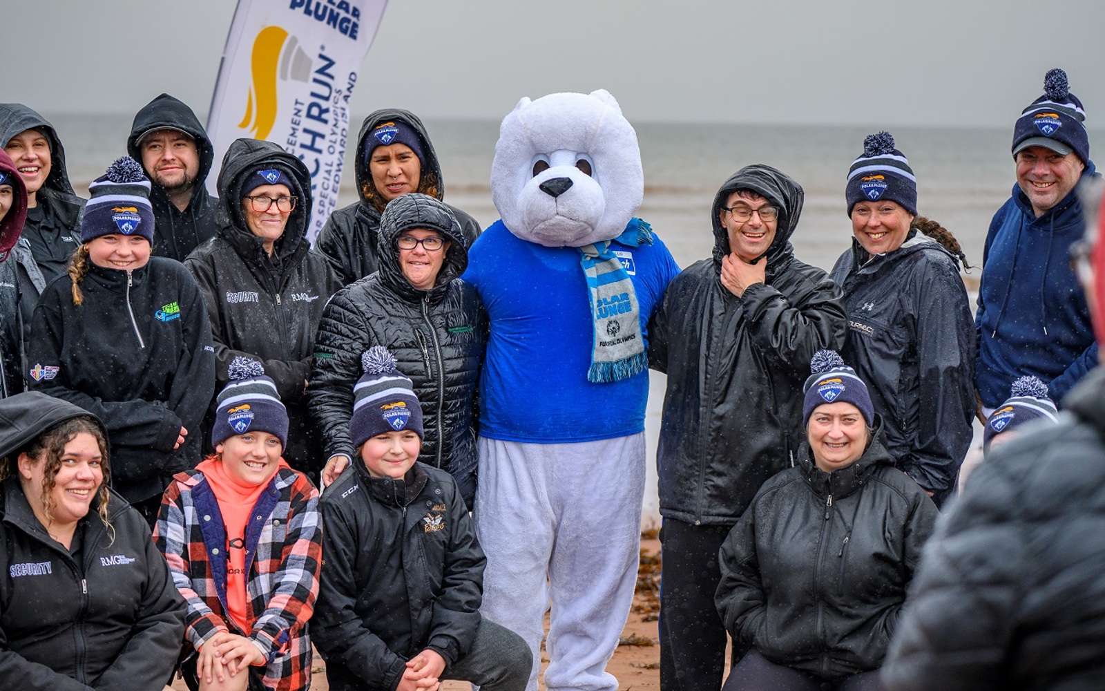 A group of people stand on a beach with a mascot after participating in a Polar Plunge