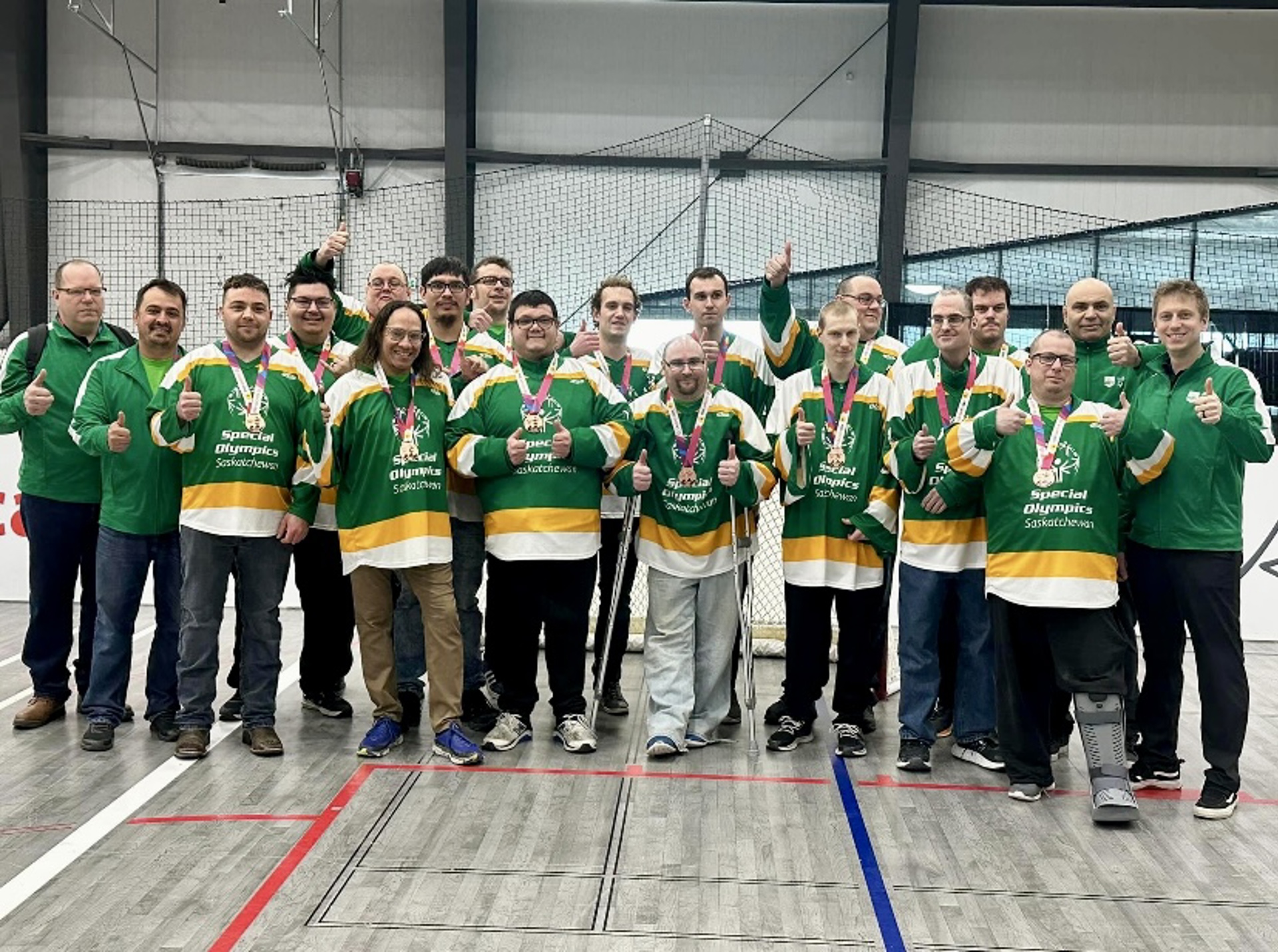 A group photo of the Special Olympics Saskatchewan Floor Hockey Team. There are 19 men in the photo, all smiling and giving thumbs up to the camera. The athletes are wearing green jerseys with medals around their necks.