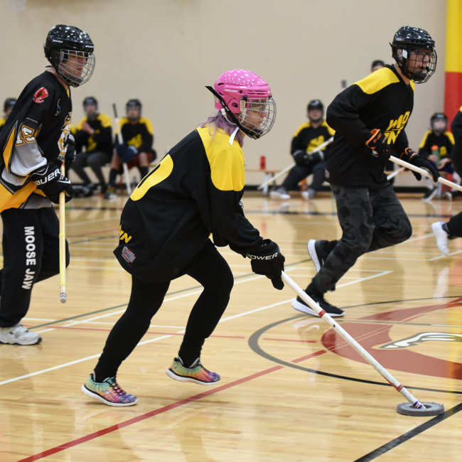 An action shot photo of athletes playing floor hockey. Four athletes can be seen running across the gym floor with their floor hockey sticks. They are all wearing helmets and black and yellow floor hockey jerseys.