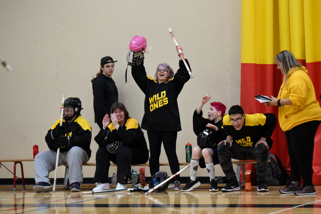 Floor hockey teammates are sitting on the bench and cheering for their teammates. Everyone is smiling, has their hands up, and are clapping for their teammates.