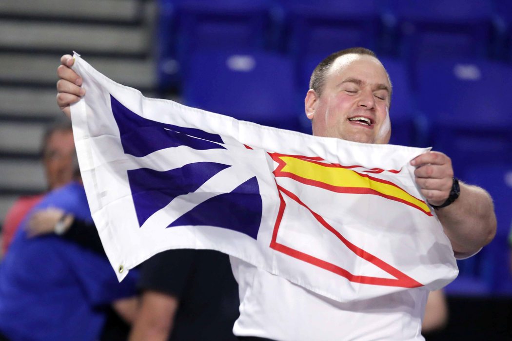 An athlete wearing a white t-shirt and thick red belt, is holding up the Newfoundland flag. He is smiling with his eyes closed. He is in clear focus as the background around him is blurred.