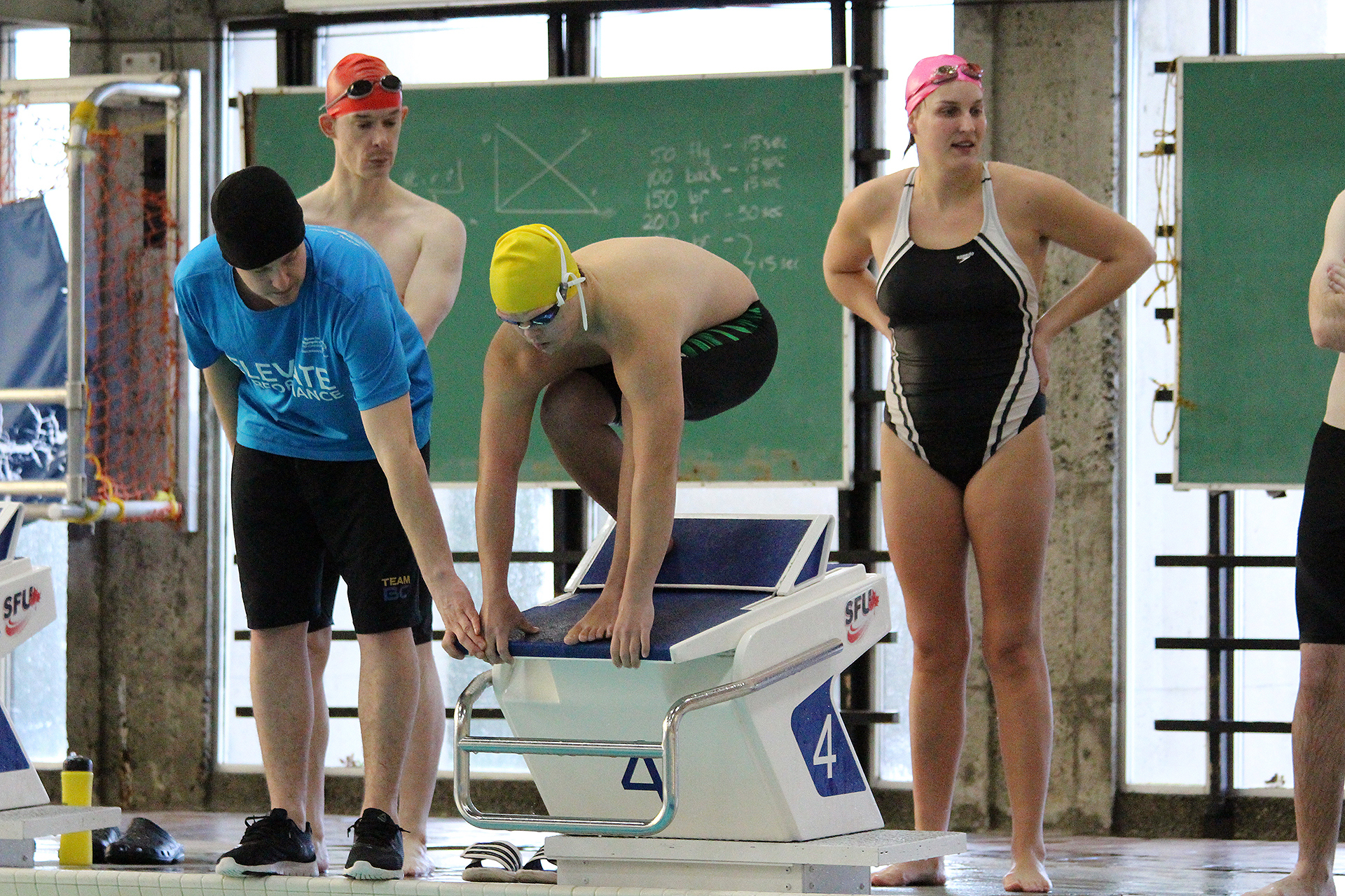 A swimmer is crouched on the starting block, waiting to go, while his team and coach surround him.