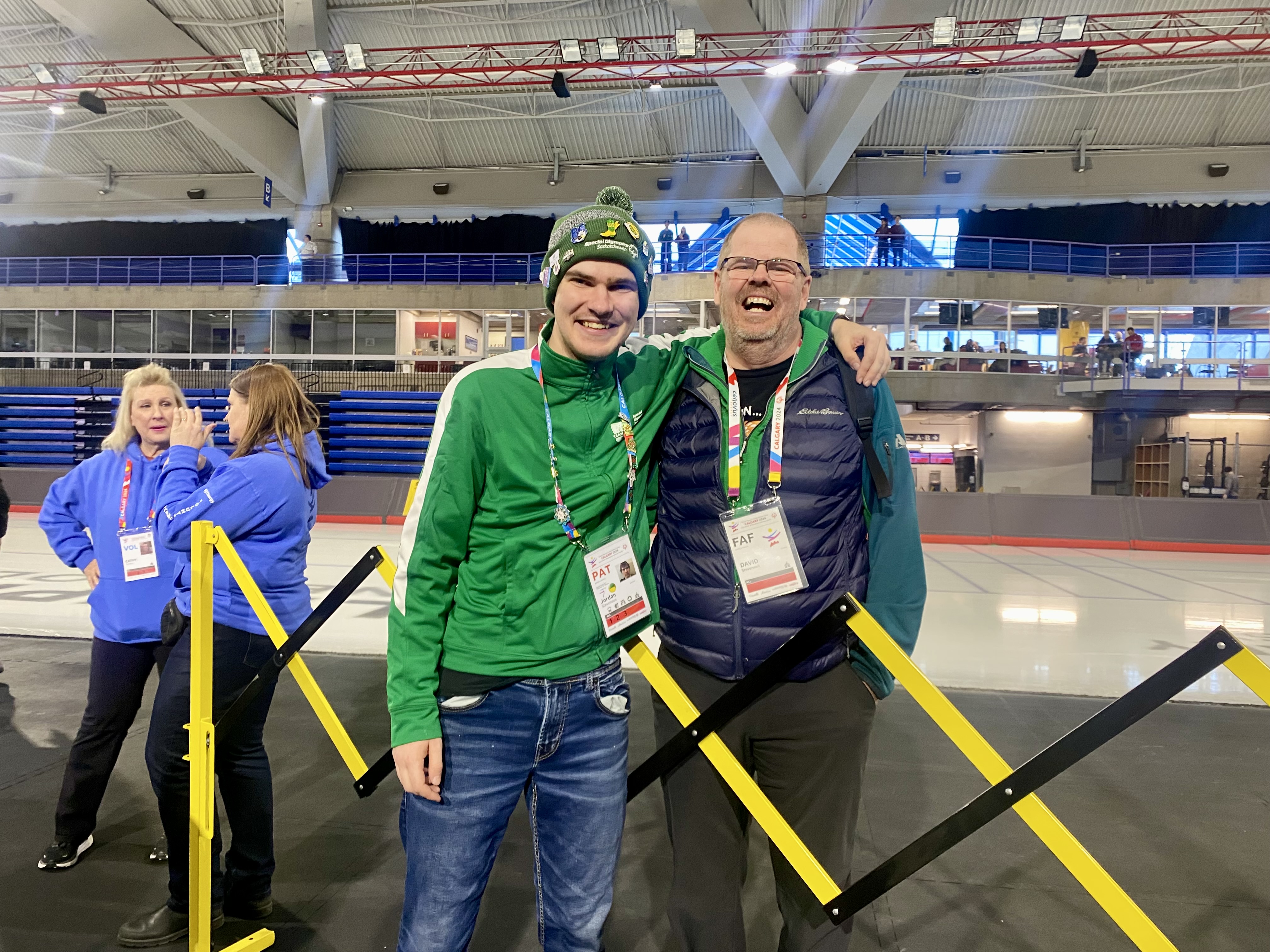 An athlete posing with his coach after the game. They are both smiling for the camera with their arms wrapped behind each other. The athlete is wearing a green sweater, a pair of jeans, and a winter hat. The coach is wearing a navy vest, and a teal sweater. They both have a lanyard around their necks. An athlete posing with his coach after the game. They are both smiling for the camera with their arms wrapped behind each other. The athlete is wearing a green sweater, a pair of jeans, and a winter hat. The coach is wearing a navy vest, and a teal sweater. They both have a lanyard around their necks.