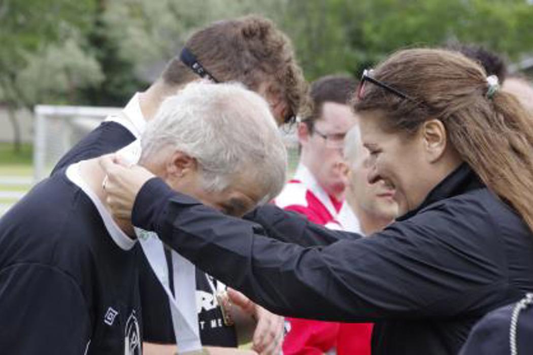 A woman on the right, wearing a black jacket, is placing a white medal around a male athlete's head. The woman is smiling and the man is bending forward so the medal could be placed around his neck.