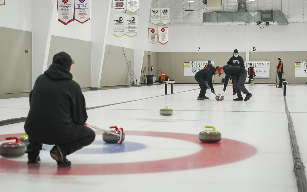 An action shot photo of a curling game. Three athletes are on the ice. One is crouching down next to the circles and the curling stones close to the camera. In the distance you can see two other athletes using their curling sticks to help the curling stone move across the ice.