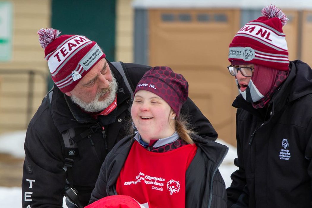 A photo of a volunteer and two athletes. They are all wearing Special Olympics Newfoundland & Labrador winter attire. The volunteer is standing behind the athletes looking around at them. The athletes are smiling.
