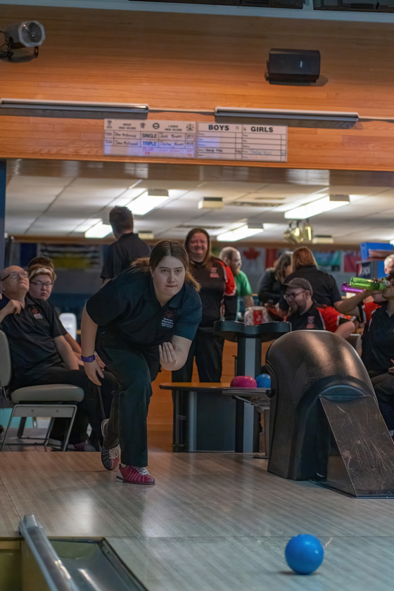 An action shot photo of a bowling athlete. She is at the start of the lane, mid throw, and you can see the blue bowling ball start to roll down the aisle. Behind the athlete, you can see her teammates smiling and watching her turn.