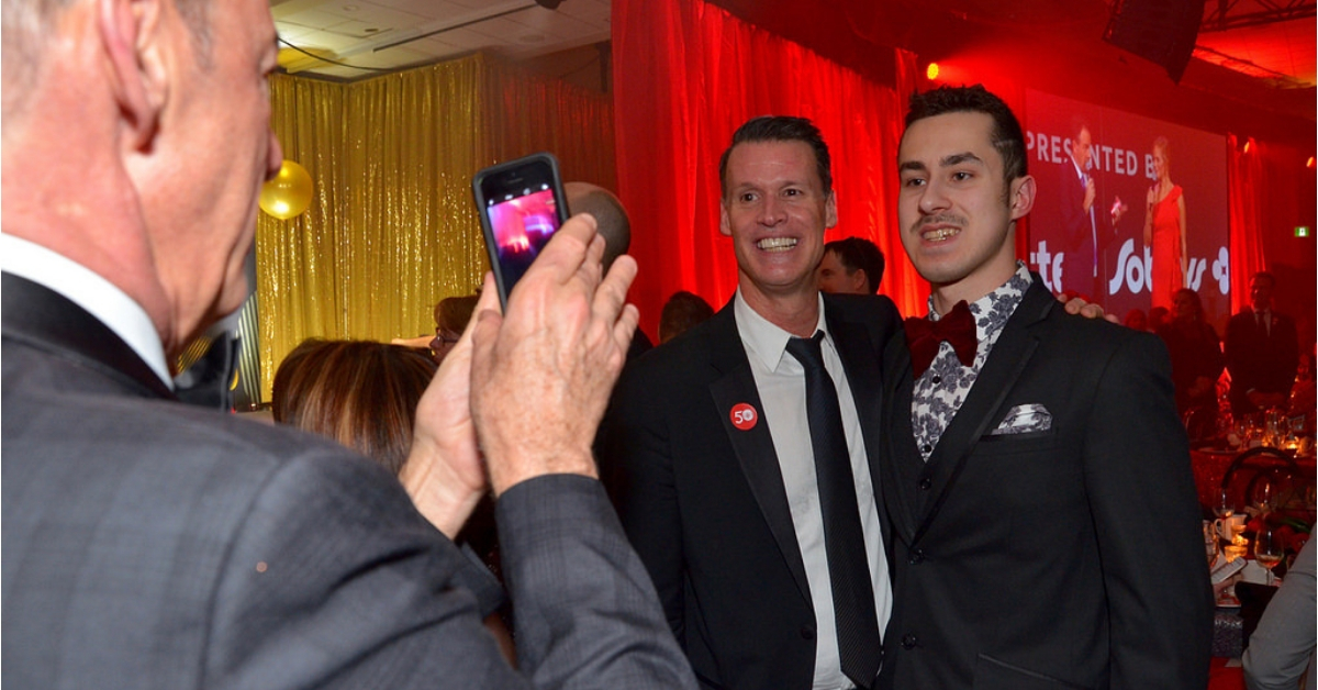 Someone takes an iPhone photo of Mark Tewksbury with a Special Olympics athlete at a gala