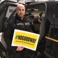 Const. Scott Edwards stands in front of a police car with his dog Chase in the front seat. Scott is holding a yellow sign that says #NoGoodWay