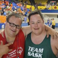 Two male Special Olympics athlete smile looking into the camera in front of a basketball court