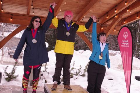 3 SOBC alpine skiing athletes on podium with hands raised and smiling with medals