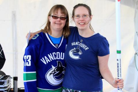 SOBC - Victoria athletes Kelsey and Jenn hugging each other with a hockey stick and wearing Canucks jerseys